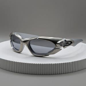 May include: Silver-toned sunglasses with a futuristic design. The sunglasses have a curved frame and dark lenses. The temples are thick and feature a unique design element. The sunglasses are displayed on a white, circular platform.