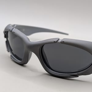May include: Gray sunglasses with dark lenses. The sunglasses have a sporty design with a thick frame and a silver emblem on the side. The lenses are dark, and the frame is a matte gray color.
