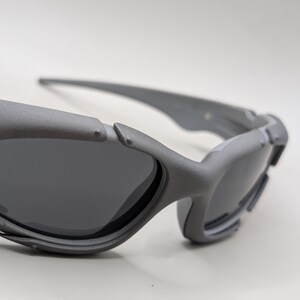 May include: Close-up of gray sunglasses with dark lenses. The sunglasses have a futuristic design with a matte finish. The arms and frame are thick and angular, with a small silver accent near the hinge.
