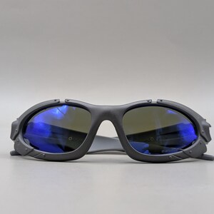 May include: Dark gray sunglasses with blue reflective lenses. The sunglasses have a thick, angular frame with a bridge and temples. The lenses are a dark color, and the frame has a matte finish. The sunglasses are designed for eye protection.