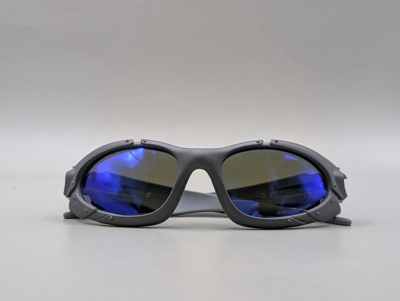 May include: Dark gray sunglasses with blue reflective lenses. The sunglasses have a thick, angular frame with a bridge and temples. The lenses are a dark color, and the frame has a matte finish. The sunglasses are designed for eye protection.