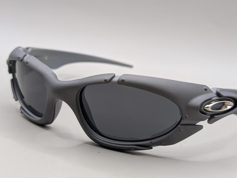 May include: Gray sunglasses with dark lenses. The sunglasses have a sporty design with a thick frame and a silver emblem on the side. The lenses are dark, and the frame is a matte gray color.