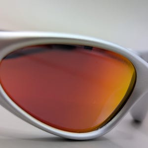May include: Close-up of silver sunglasses with a curved frame and orange-red lenses. The lenses transition from a deep red to a vibrant orange hue. The Oakley logo is visible on the temple.