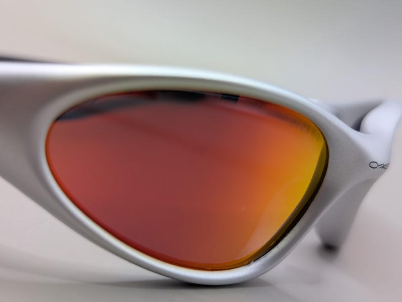 May include: Close-up of silver sunglasses with a curved frame and orange-red lenses. The lenses transition from a deep red to a vibrant orange hue. The Oakley logo is visible on the temple.