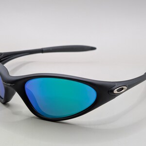 May include: Black sunglasses with blue and green mirrored lenses. The sunglasses have a curved frame and a small silver logo on the side. The lenses reflect a gradient of blue and green.
