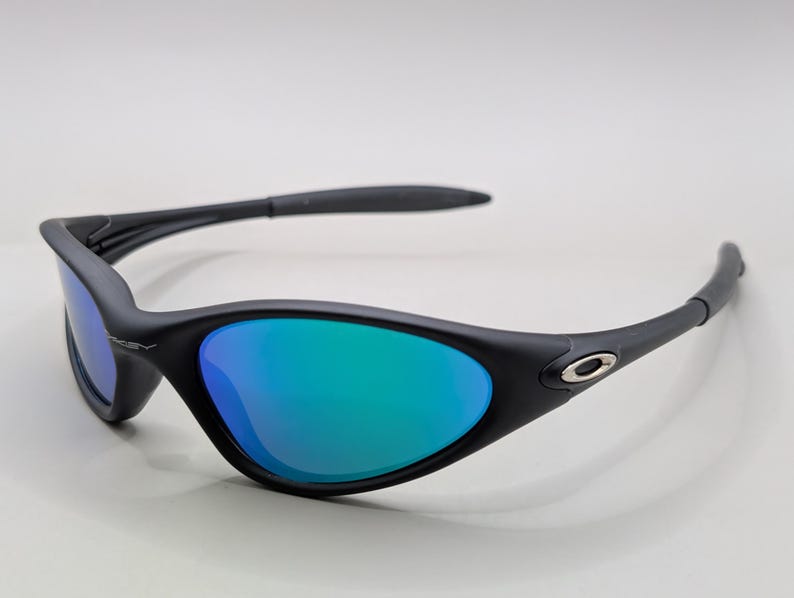 May include: Black sunglasses with blue and green mirrored lenses. The sunglasses have a curved frame and a small silver logo on the side. The lenses reflect a gradient of blue and green.