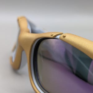 K&ouml;nnte beinhalten: Nahaufnahme einer Sonnenbrille mit goldfarbenen Rahmen und violett get&ouml;nten Gl&auml;sern. Die Rahmen haben einen wei&szlig;en Akzent auf der Innenseite. Die Sonnenbrille ist mit einer geschwungenen Form und einem Scharnierdetail versehen.