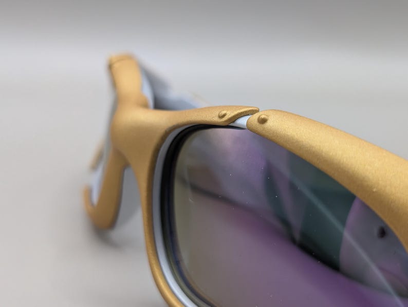 K&ouml;nnte beinhalten: Nahaufnahme einer Sonnenbrille mit goldfarbenen Rahmen und violett get&ouml;nten Gl&auml;sern. Die Rahmen haben einen wei&szlig;en Akzent auf der Innenseite. Die Sonnenbrille ist mit einer geschwungenen Form und einem Scharnierdetail versehen.