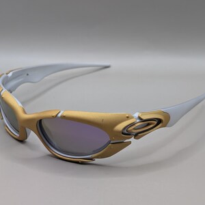 K&ouml;nnte beinhalten: Goldene und wei&szlig;e Sportsonnenbrille. Die Fassung ist eine Kombination aus Gold und Wei&szlig;, mit einer geschwungenen Form und verspiegelten Gl&auml;sern. Die Sonnenbrille ist f&uuml;r Sport und Outdoor-Aktivit&auml;ten konzipiert.