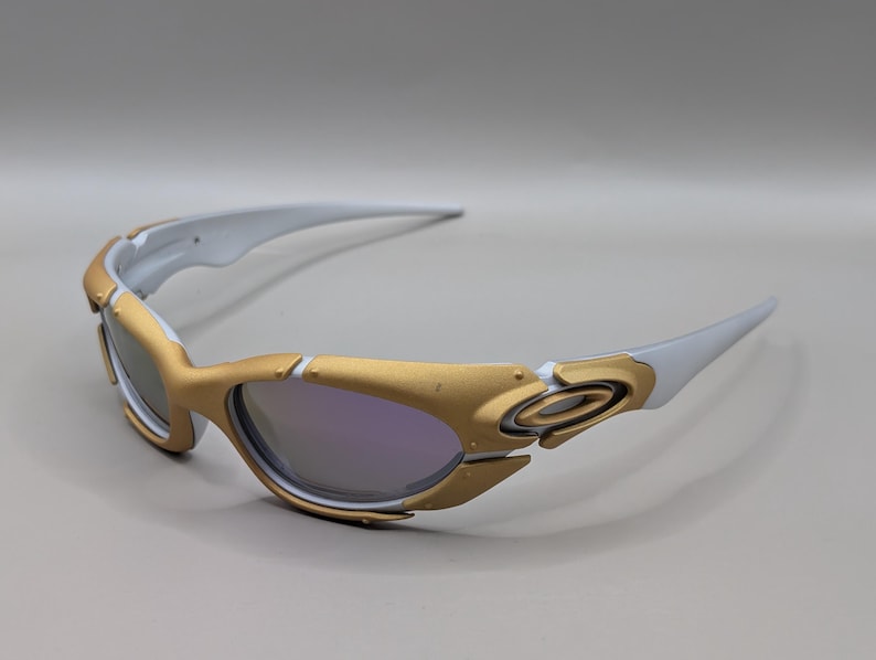 K&ouml;nnte beinhalten: Goldene und wei&szlig;e Sportsonnenbrille. Die Fassung ist eine Kombination aus Gold und Wei&szlig;, mit einer geschwungenen Form und verspiegelten Gl&auml;sern. Die Sonnenbrille ist f&uuml;r Sport und Outdoor-Aktivit&auml;ten konzipiert.