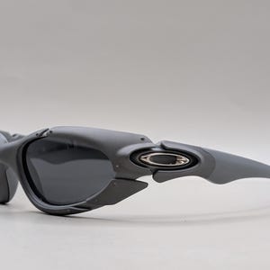 May include: Gray sunglasses with dark lenses. The sunglasses have a curved design with a silver emblem on the temple. The frame is a matte gray color.