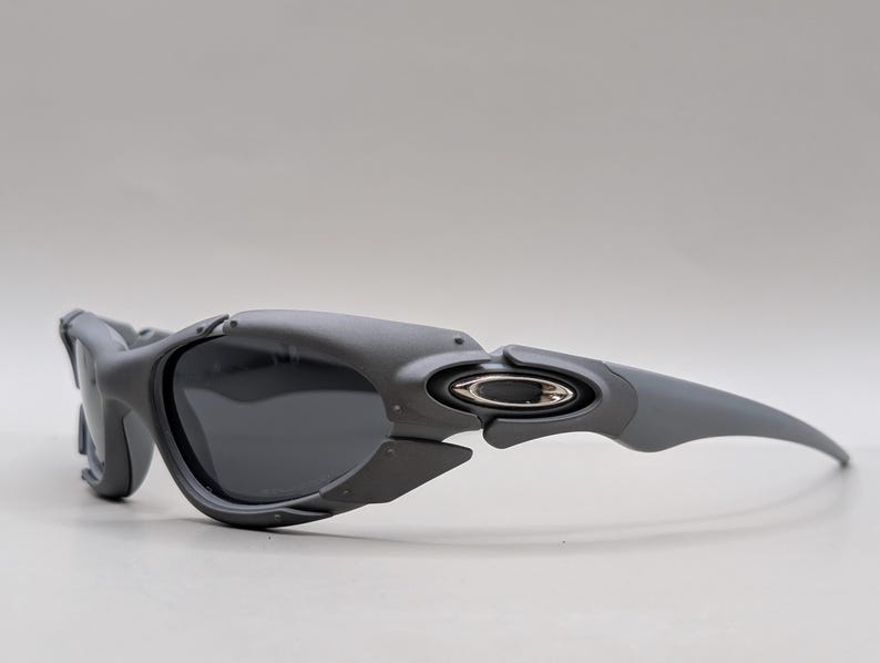 May include: Gray sunglasses with dark lenses. The sunglasses have a curved design with a silver emblem on the temple. The frame is a matte gray color.