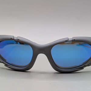 K&ouml;nnte beinhalten: Graue Sportsonnenbrille mit blau verspiegelten Gl&auml;sern. Die Sonnenbrille hat ein Wrap-around-Design mit mattem Finish. Die Gl&auml;ser reflektieren eine leuchtend blaue Farbe, und der Rahmen hat eine einzigartige, eckige Form.