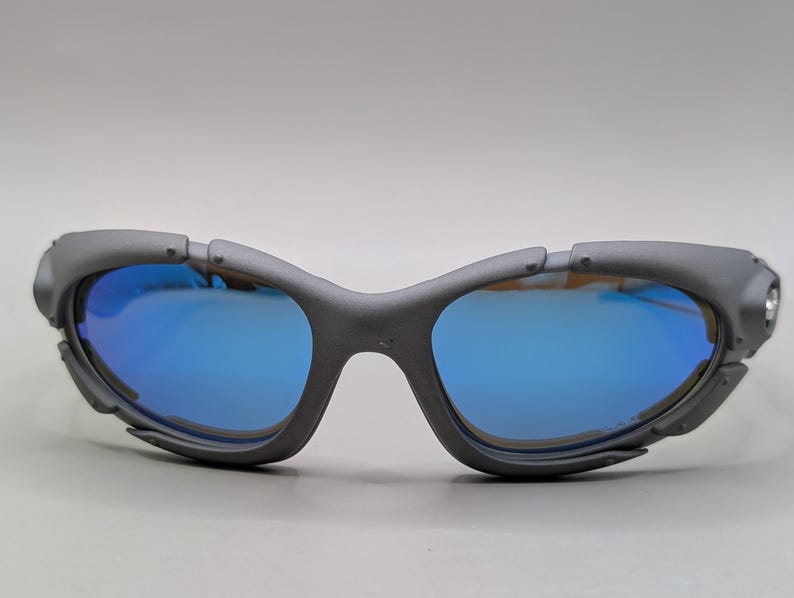 K&ouml;nnte beinhalten: Graue Sportsonnenbrille mit blau verspiegelten Gl&auml;sern. Die Sonnenbrille hat ein Wrap-around-Design mit mattem Finish. Die Gl&auml;ser reflektieren eine leuchtend blaue Farbe, und der Rahmen hat eine einzigartige, eckige Form.