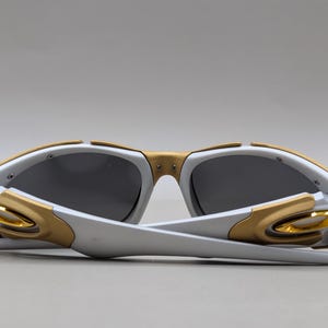 May include: White and gold sunglasses with dark gray lenses. The sunglasses have a modern design with gold accents on the frame and temples. The arms are white and fold inward. The lenses are a dark gray color.