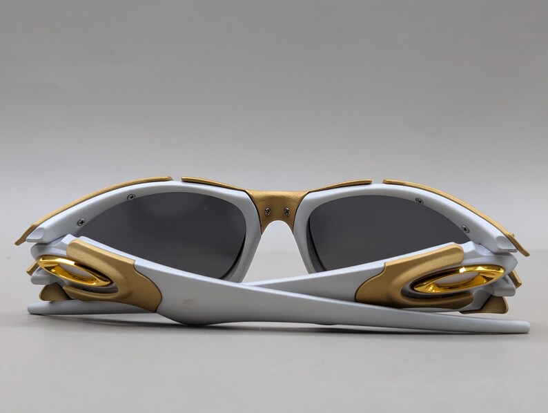 May include: White and gold sunglasses with dark gray lenses. The sunglasses have a modern design with gold accents on the frame and temples. The arms are white and fold inward. The lenses are a dark gray color.