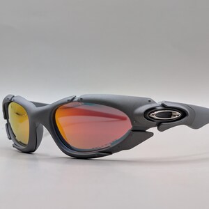 Puede incluir: Gafas de sol deportivas grises con lentes rojas y amarillas. Las gafas tienen un dise&ntilde;o envolvente con una montura gris oscuro y el logotipo de Oakley en las patillas. Las lentes tienen un acabado espejado.
