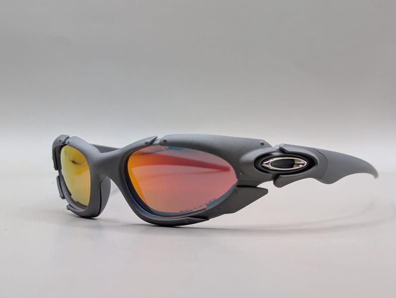 Puede incluir: Gafas de sol deportivas grises con lentes rojas y amarillas. Las gafas tienen un dise&ntilde;o envolvente con una montura gris oscuro y el logotipo de Oakley en las patillas. Las lentes tienen un acabado espejado.