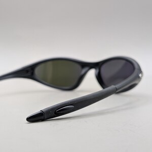 May include: Black sunglasses with dark lenses. The sunglasses have a sleek, curved design with a matte finish. The temples are extended and angled, and the lenses are a dark gray color.