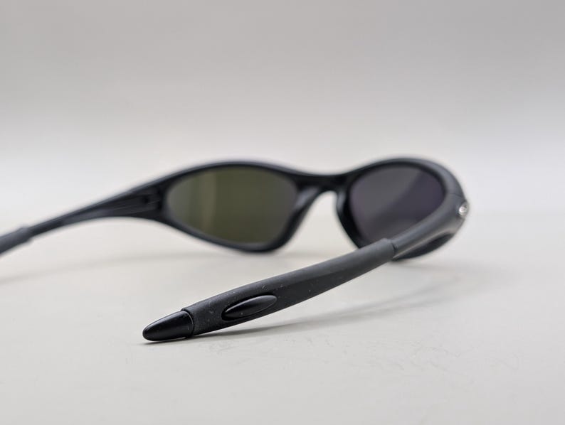 May include: Black sunglasses with dark lenses. The sunglasses have a sleek, curved design with a matte finish. The temples are extended and angled, and the lenses are a dark gray color.