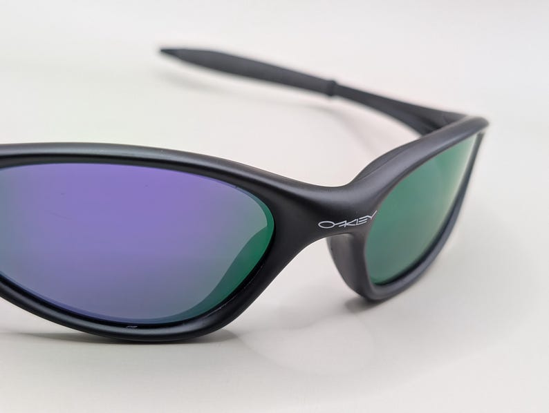 May include: Close-up of black sunglasses with purple and green lenses. The sunglasses have a sleek, modern design with the brand name "Oakley" printed on the side. The lenses reflect a gradient of colors.