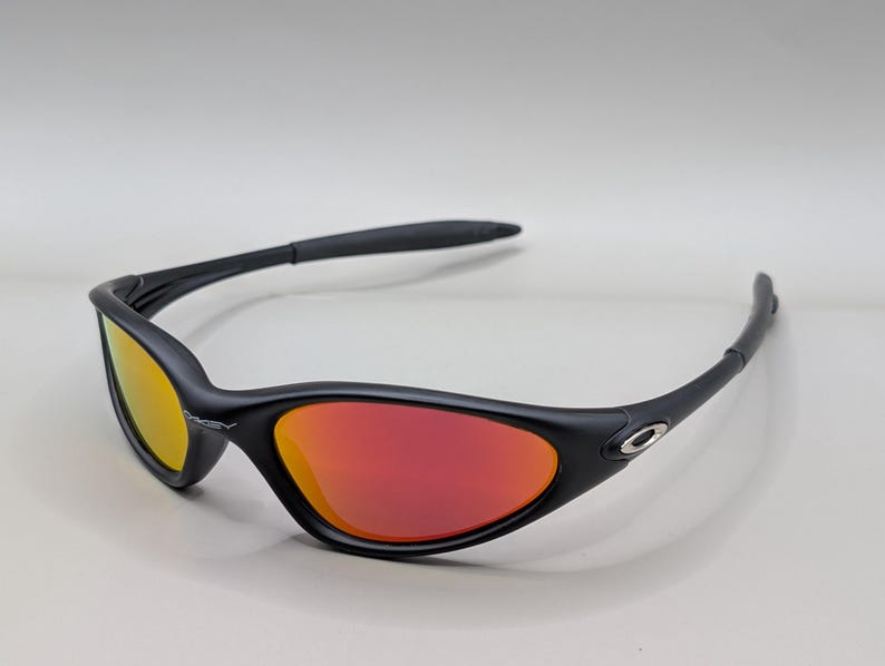 Pode incluir: &Oacute;culos de sol pretos com arma&ccedil;&atilde;o curva e lentes vermelhas e amarelas. Os &oacute;culos de sol t&ecirc;m um design elegante e moderno, com um pequeno logotipo prateado na lateral da arma&ccedil;&atilde;o. As lentes fazem uma transi&ccedil;&atilde;o do amarelo para o vermelho.