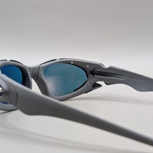 Puede incluir: Gafas de sol grises con lentes azules. Las gafas de sol tienen un dise&ntilde;o moderno con una montura y patillas curvas. El texto "MADE IN U.S.A." es visible en la patilla.