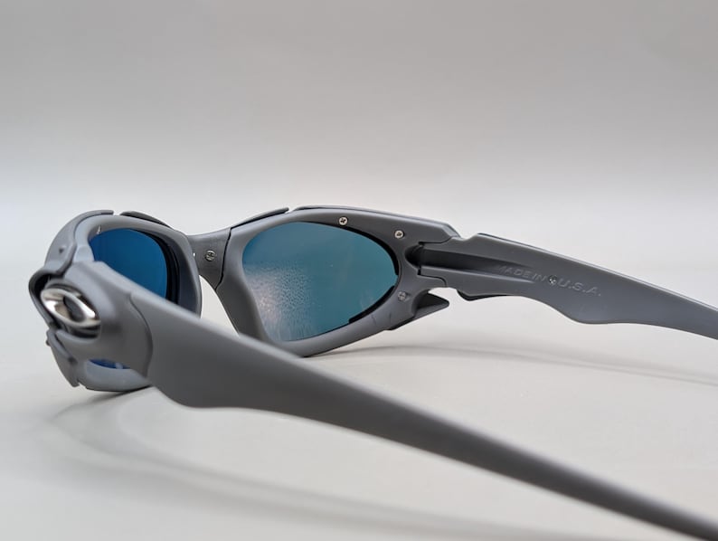 Puede incluir: Gafas de sol grises con lentes azules. Las gafas de sol tienen un dise&ntilde;o moderno con una montura y patillas curvas. El texto "MADE IN U.S.A." es visible en la patilla.
