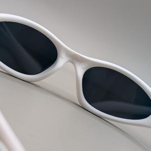 May include: White sunglasses with dark lenses. The sunglasses have a curved, streamlined design with a thick frame. The lenses are oval-shaped and appear to be tinted. The arms of the sunglasses have a textured design.