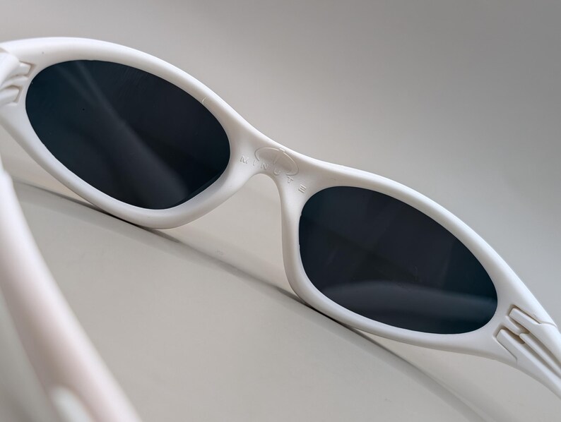 May include: White sunglasses with dark lenses. The sunglasses have a curved, streamlined design with a thick frame. The lenses are oval-shaped and appear to be tinted. The arms of the sunglasses have a textured design.