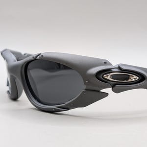 May include: Gray sunglasses with dark lenses. The sunglasses have a unique, angular design with a matte finish. The temples feature a silver emblem. The overall aesthetic is modern and sporty.