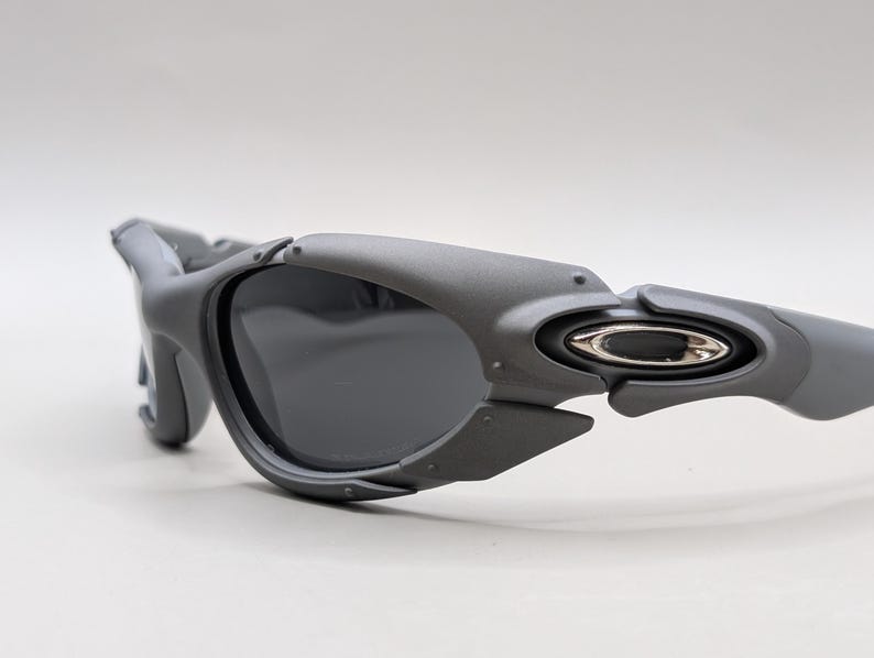 May include: Gray sunglasses with dark lenses. The sunglasses have a unique, angular design with a matte finish. The temples feature a silver emblem. The overall aesthetic is modern and sporty.