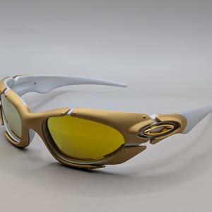 Pu&ograve; includere: Occhiali da sole dorati e bianchi con lenti gialle. Gli occhiali da sole hanno un design sportivo con montatura dorata e aste bianche. Le lenti sono di colore giallo brillante e lo stile generale &egrave; moderno ed elegante.