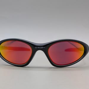 Pode incluir: &Oacute;culos de sol pretos com lentes degrad&ecirc; vermelhas e laranjas. Os &oacute;culos de sol t&ecirc;m um formato oval curvo e a marca "OAKLEY" est&aacute; impressa na arma&ccedil;&atilde;o.