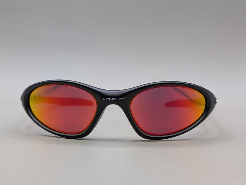 Pode incluir: &Oacute;culos de sol pretos com lentes degrad&ecirc; vermelhas e laranjas. Os &oacute;culos de sol t&ecirc;m um formato oval curvo e a marca "OAKLEY" est&aacute; impressa na arma&ccedil;&atilde;o.