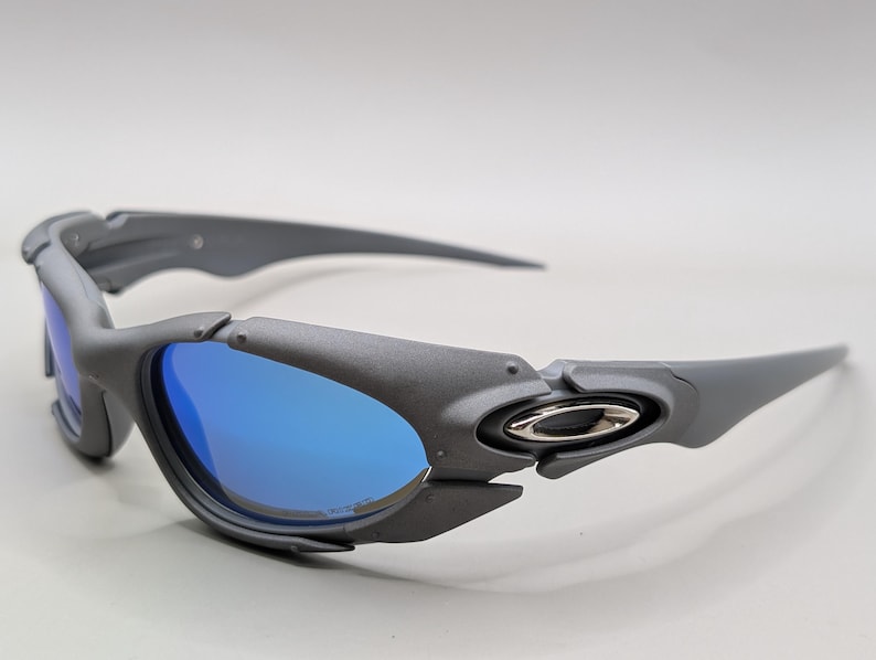 K&ouml;nnte beinhalten: Graue Sonnenbrille mit blauen Gl&auml;sern. Die Sonnenbrille hat ein sportliches Design mit matter Oberfl&auml;che. Die Gl&auml;ser sind leuchtend blau und die Rahmen sind ein ged&auml;mpftes Grau. Die B&uuml;gel der Sonnenbrille sind lang und schlank.