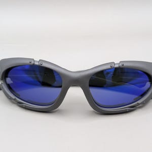 May include: Gray sunglasses with blue lenses. The sunglasses have a unique, angular frame design with visible rivets and a futuristic aesthetic. The lenses are a vibrant blue, contrasting with the gray frame.
