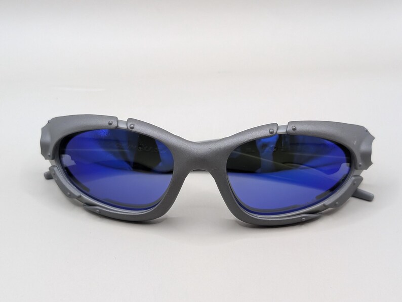 May include: Gray sunglasses with blue lenses. The sunglasses have a unique, angular frame design with visible rivets and a futuristic aesthetic. The lenses are a vibrant blue, contrasting with the gray frame.