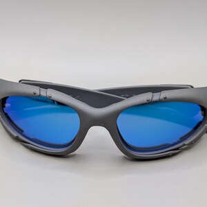 K&ouml;nnte beinhalten: Graue Sonnenbrille mit blauen Gl&auml;sern. Die Sonnenbrille im Wickeldesign hat eine matte Oberfl&auml;che. Die Gl&auml;ser sind leuchtend blau und kontrastieren mit dem grauen Rahmen. Die B&uuml;gel haben eine einzigartige, eckige Form.