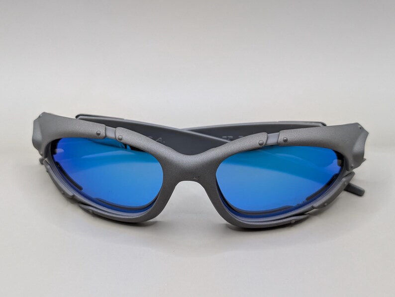 K&ouml;nnte beinhalten: Graue Sonnenbrille mit blauen Gl&auml;sern. Die Sonnenbrille im Wickeldesign hat eine matte Oberfl&auml;che. Die Gl&auml;ser sind leuchtend blau und kontrastieren mit dem grauen Rahmen. Die B&uuml;gel haben eine einzigartige, eckige Form.