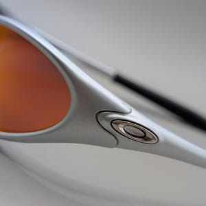 May include: Close-up of silver sunglasses with orange and yellow lenses. The sunglasses have a sleek, modern design with a metallic finish. The lenses are a gradient of colors, transitioning from orange to yellow.