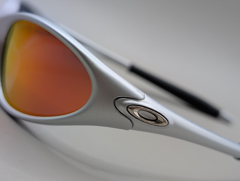 May include: Close-up of silver sunglasses with orange and yellow lenses. The sunglasses have a sleek, modern design with a metallic finish. The lenses are a gradient of colors, transitioning from orange to yellow.