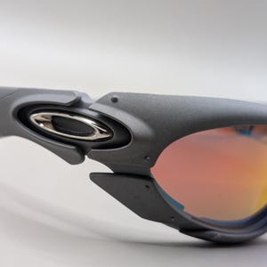 Puede incluir: Primer plano de gafas de sol grises con un emblema plateado en la patilla. Las lentes tienen un degradado de naranja y amarillo. Las gafas de sol tienen un dise&ntilde;o futurista.