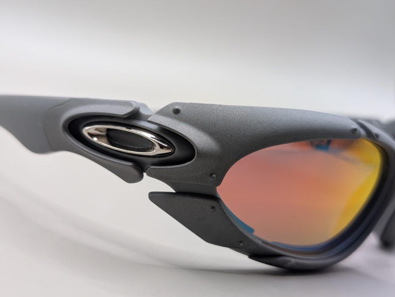 Puede incluir: Primer plano de gafas de sol grises con un emblema plateado en la patilla. Las lentes tienen un degradado de naranja y amarillo. Las gafas de sol tienen un dise&ntilde;o futurista.