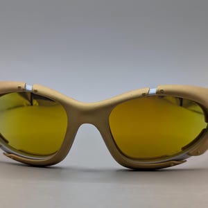 Pu&ograve; includere: Occhiali da sole color oro con lenti gialle. La montatura presenta un design futuristico unico con dettagli argentati e un piccolo dettaglio circolare sul lato. Le lenti sono di un giallo brillante e lo stile generale &egrave; sportivo e moderno.