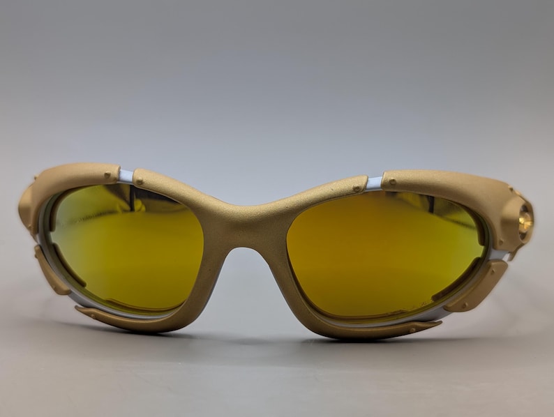 Pu&ograve; includere: Occhiali da sole color oro con lenti gialle. La montatura presenta un design futuristico unico con dettagli argentati e un piccolo dettaglio circolare sul lato. Le lenti sono di un giallo brillante e lo stile generale &egrave; sportivo e moderno.