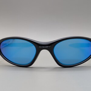 K&ouml;nnte beinhalten: Schwarze Sonnenbrille mit blau verspiegelten Gl&auml;sern. Die Sonnenbrille hat eine geschwungene, ovale Form und der Markenname "OAKLEY" ist wei&szlig; auf der Br&uuml;cke aufgedruckt.