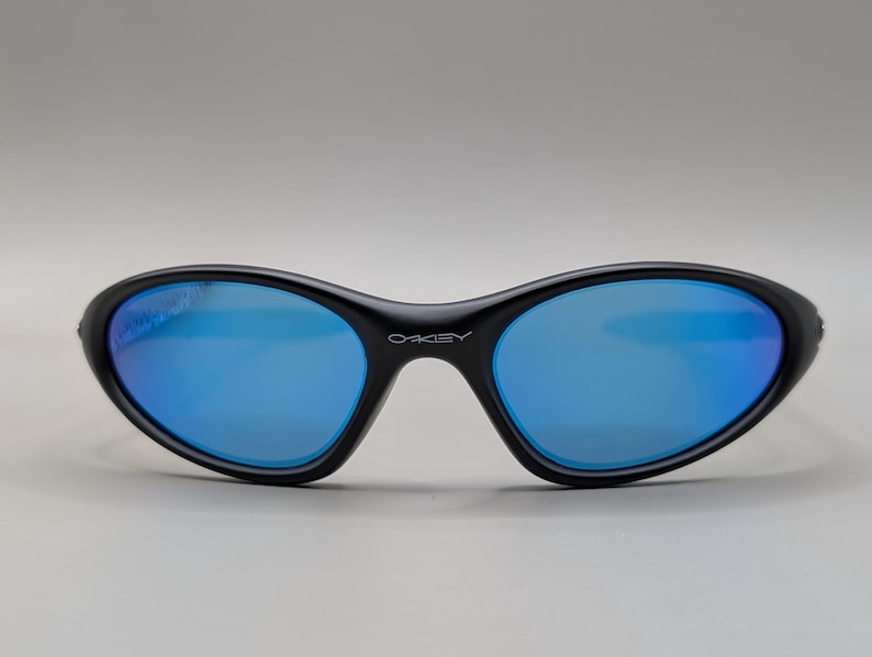 K&ouml;nnte beinhalten: Schwarze Sonnenbrille mit blau verspiegelten Gl&auml;sern. Die Sonnenbrille hat eine geschwungene, ovale Form und der Markenname "OAKLEY" ist wei&szlig; auf der Br&uuml;cke aufgedruckt.