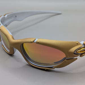May include: Gold and white sunglasses with a reflective lens. The sunglasses have a gold frame with white accents on the arms. The lenses have a gradient of orange and yellow.