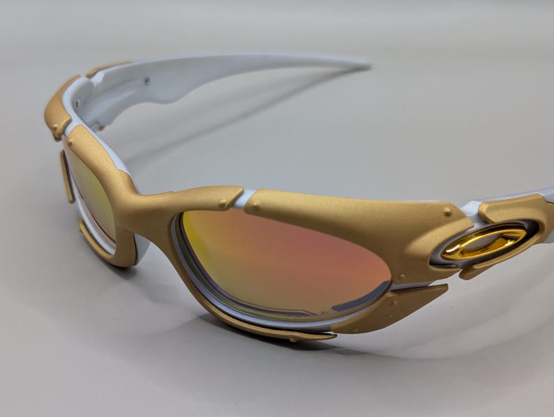 May include: Gold and white sunglasses with a reflective lens. The sunglasses have a gold frame with white accents on the arms. The lenses have a gradient of orange and yellow.